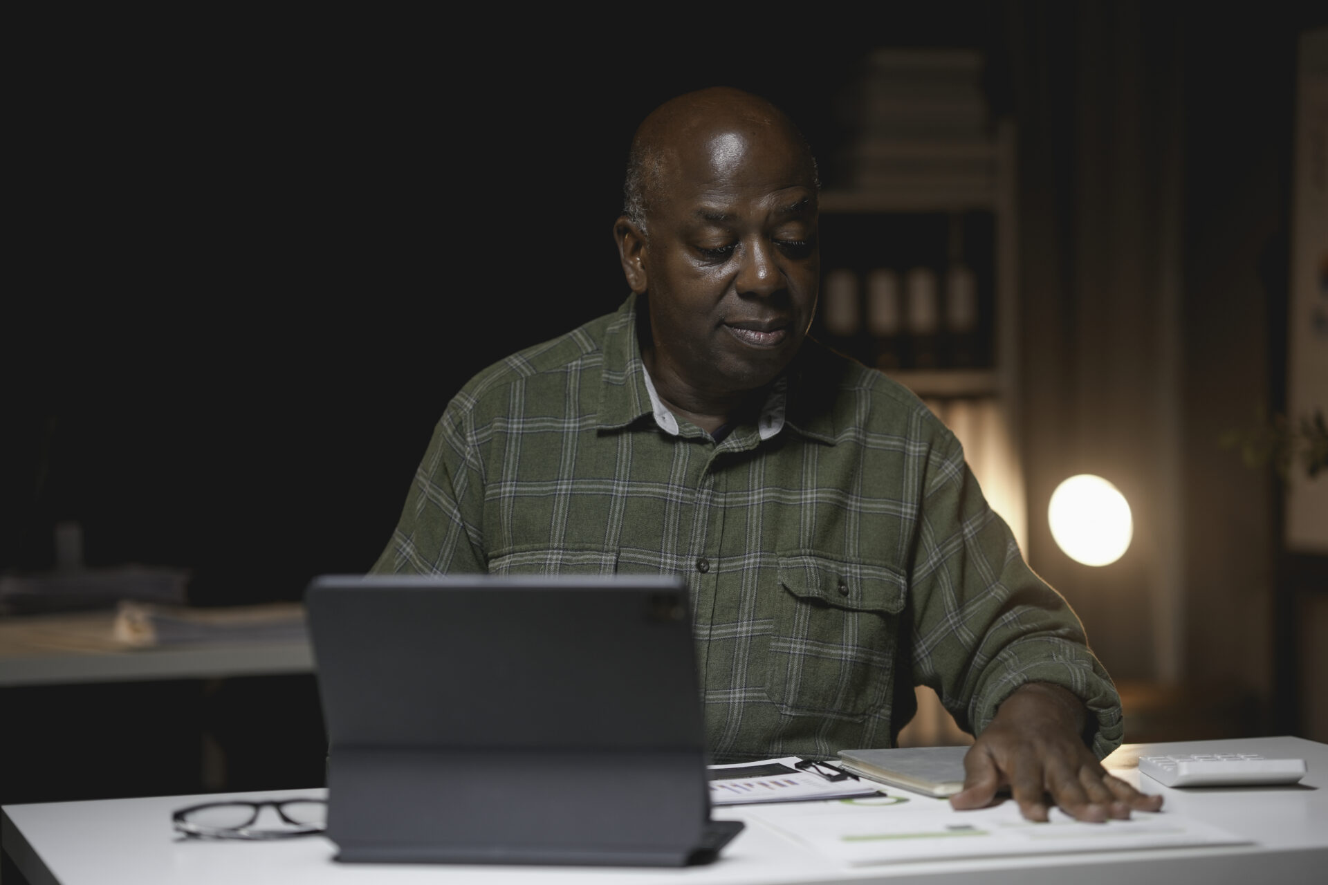 Mature man focuses intently on a digital tablet while working late at his office desk. The warm lighting and organized workspace create a productive atmosphere