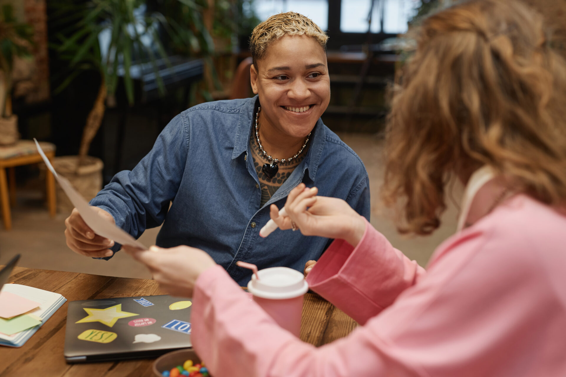 Portrait of smiling masc Black woman discussing project with colleague at meeting table and holding documents