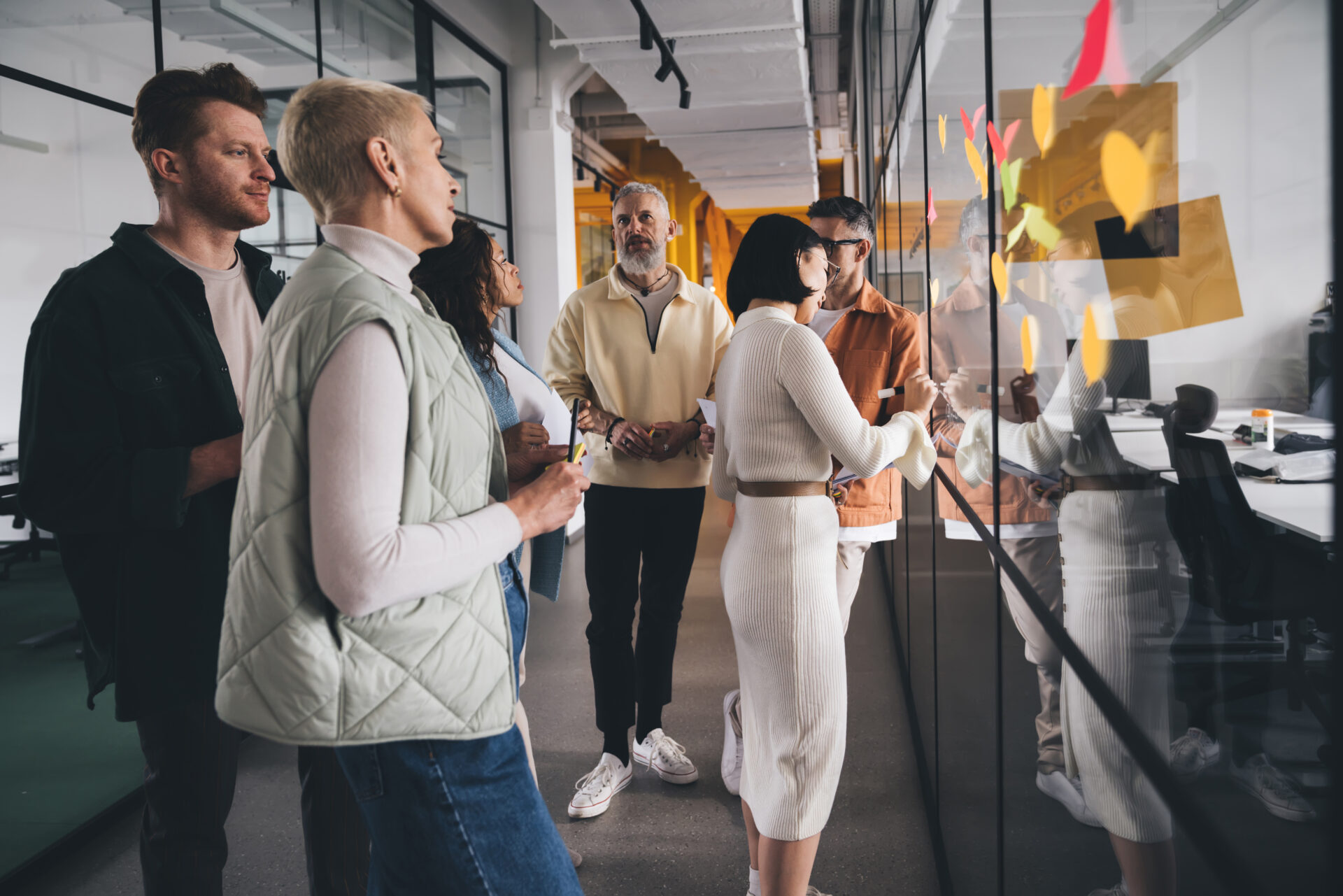 Group of diverse people standing near glass wall with colorful sticky notes in workplace and discussing ideas of startup project during brainstorm