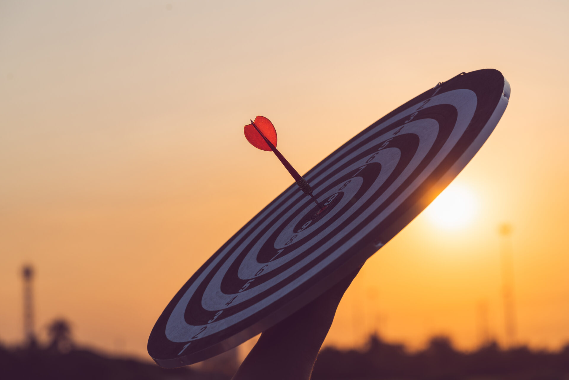 Close up red dart arrow hitting target center dartboard on sunset background. Business targeting and focus concept