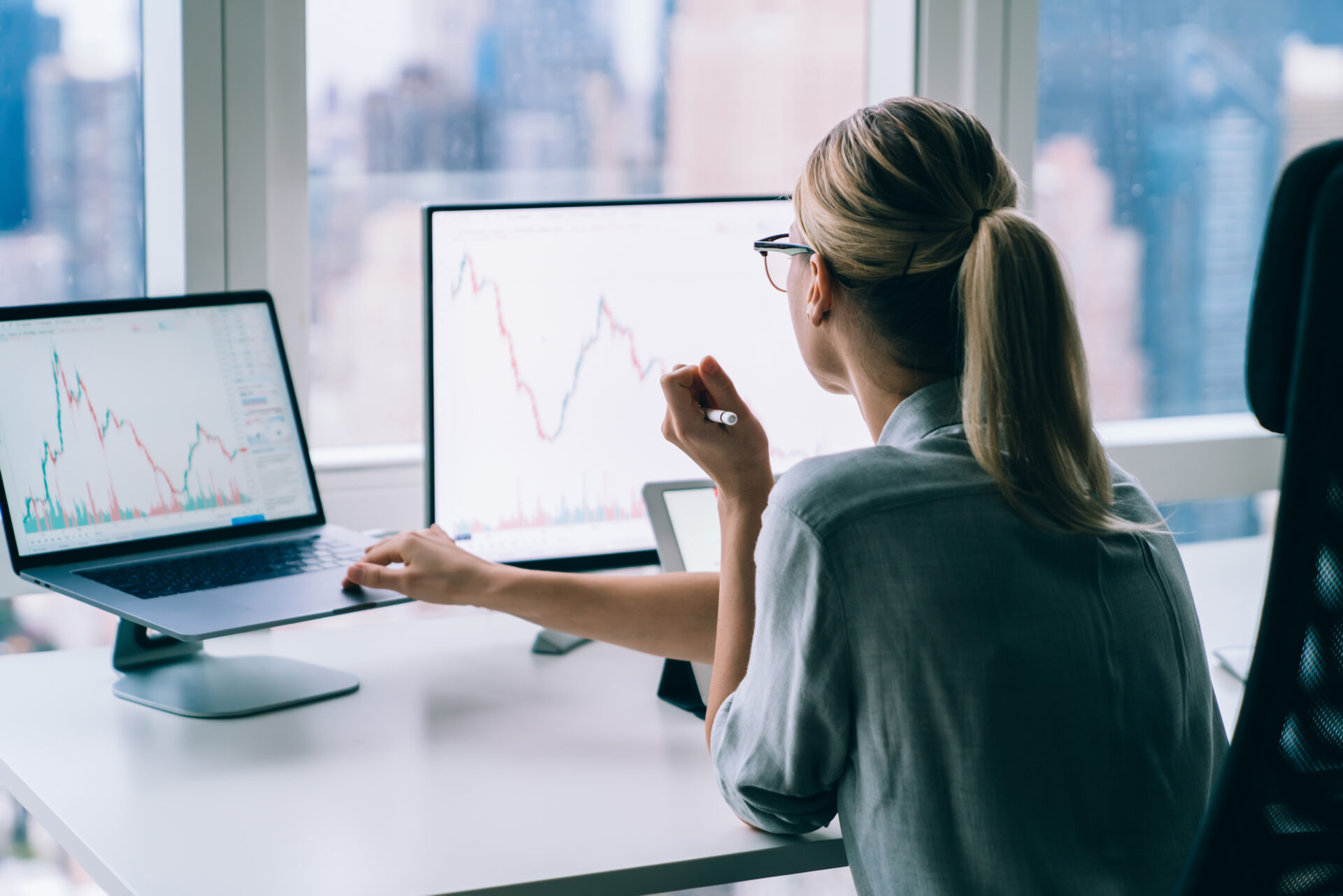 Back view of concentrated lady in formal wear and eyeglasses using laptop while working on project and analyzing diagrams in workplace