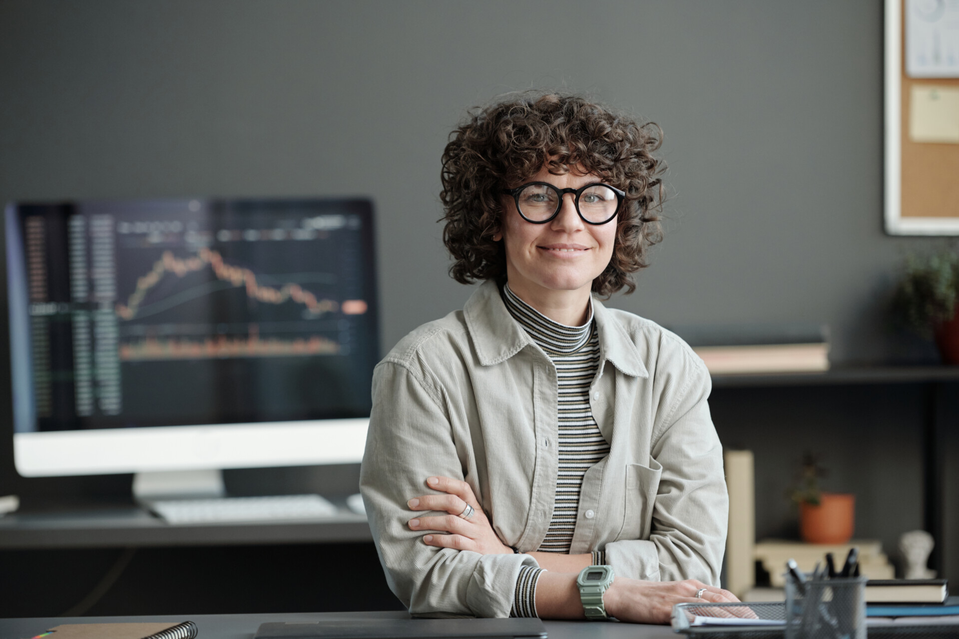 Young curly brunette businesswoman in casualwear and eyeglasses sitting by workplace against computer screen with graph and chart